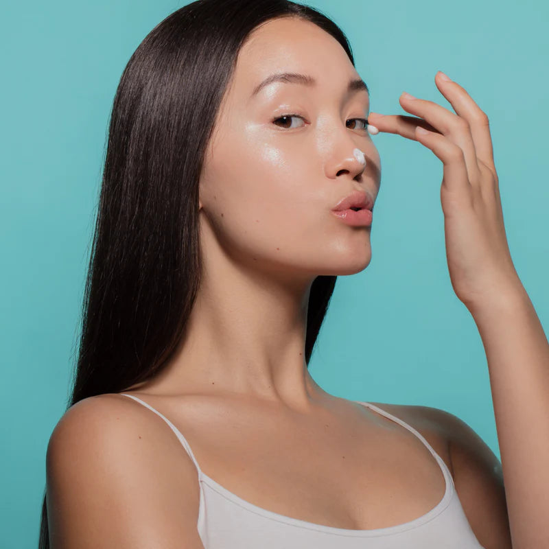 Woman with dark hair applying white cream to her nose and eye, making a kissing expression.
