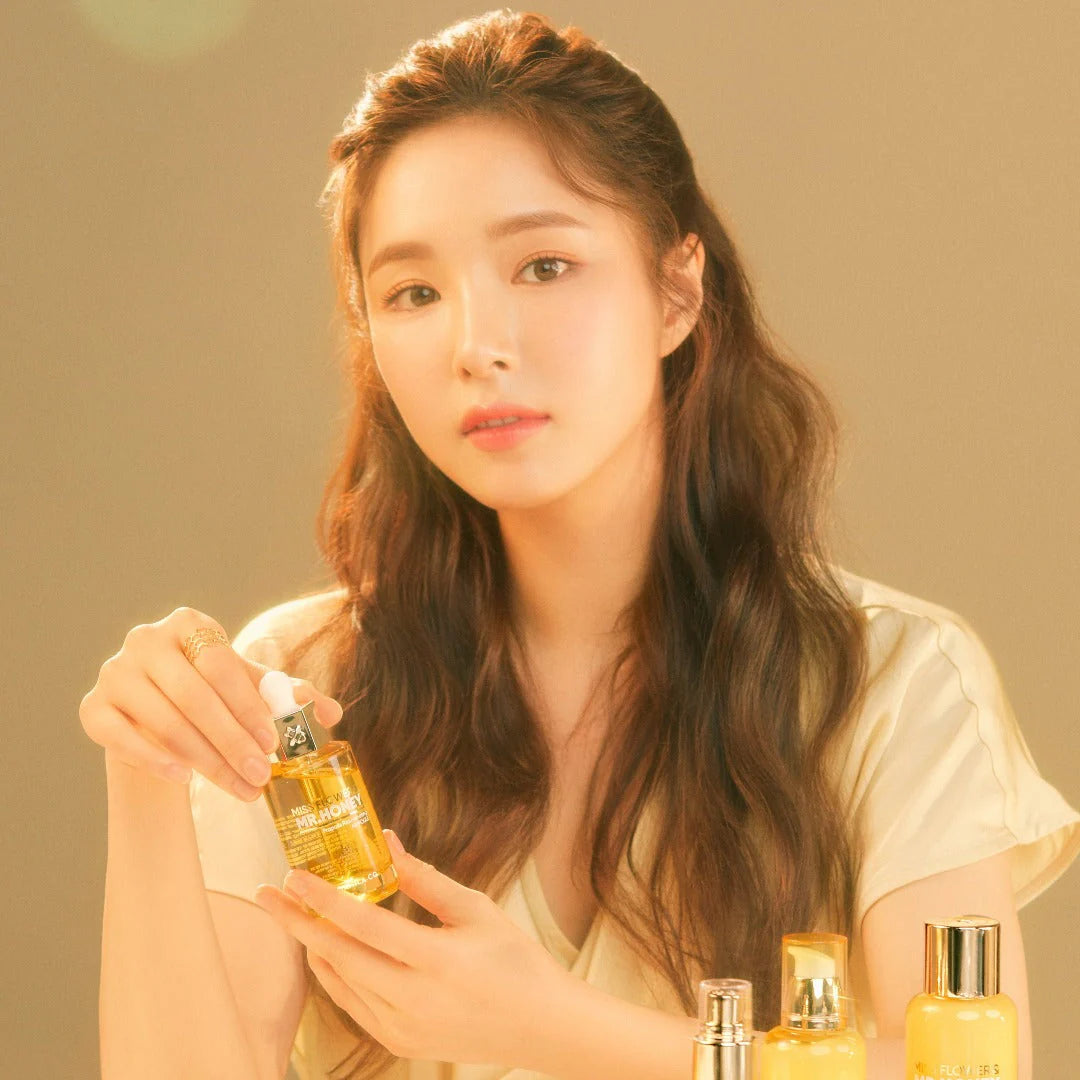 Woman holding a "MISS FLOWERS & MR. HONEY" skincare serum, with other beauty products on a warm-toned background.