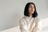 Young woman with short dark hair in a light sweater, looking thoughtfully to the right against a pale wall.