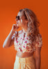 Blonde woman in sunglasses and a floral crop top, hand to chin, looking right against an orange wall.